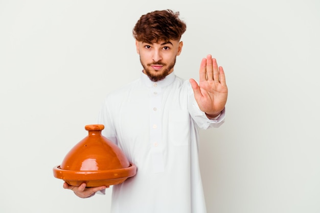 Junger marokkanischer Mann, der das typische arabische Kostüm hält, das eine Tajine lokalisiert auf weißem Stehen mit ausgestreckter Hand hält, die Stoppschild zeigt, das Sie verhindert.