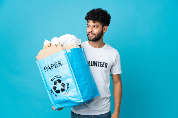 Junger marokkanischer Mann, der einen Recyclingbeutel voll Papier hält, um über isolierte Wand zu recyceln, die zur Seite schaut und lächelt