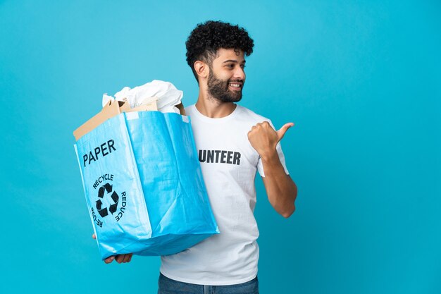 Junger marokkanischer Mann, der einen Recyclingbeutel voll Papier hält, um über isolierte Wand zu recyceln, die zur Seite zeigt, um ein Produkt zu präsentieren