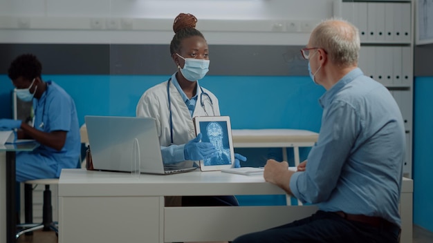 Junger Mediziner, der einem älteren kranken Mann in einer Gesundheitseinrichtung mit Plexiglaswand moderne Tablette mit Röntgenscan zeigt. Arzt, der das Röntgenergebnis auf einem digitalen Gerät für eine professionelle Untersuchung hält