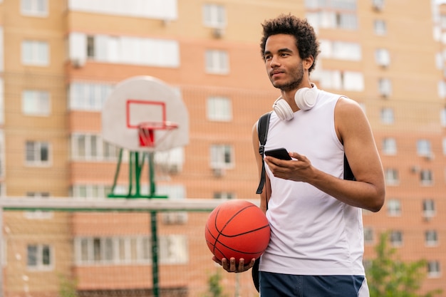 Junger Mischrassensportler mit Ball- und Smartphone-SMS in der Pause auf dem Spielplatz in der städtischen Umgebung