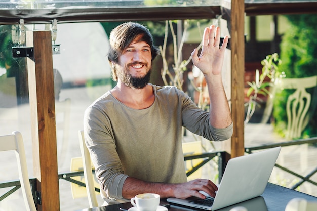 Junger moderner Mann, der im Café mit dem Laptop wellenartig bewegt seine Hand sitzt.