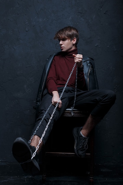 Junger moderner Mann in einer modischen Jacke im Vintage-Burgundergolf in trendigen Jeans und schwarzen Turnschuhen mit einer Metallkette sitzt auf einem Holzstuhl in einem Raum nahe einer schwarzen Wand. Hübscher stilvoller Kerl