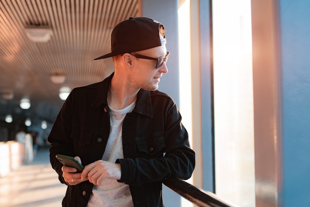 Junger modischer moderner Kerl, ein Mann mit schwarzer Mütze und Sonnenbrille mit Smartphone, telefoniert auf einem städtischen Hintergrund in einem Tunnel in den Sonnenuntergangsstrahlen der Sonne.