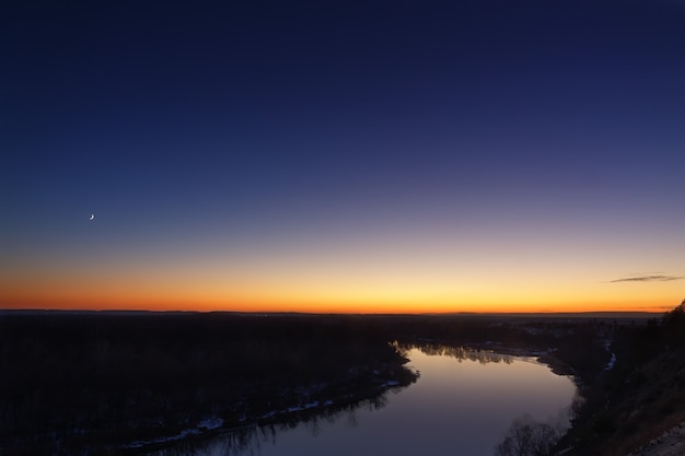 Junger Mond über dem Fluss in der Abenddämmerung. Heller Abendhimmel nach Sonnenuntergang.