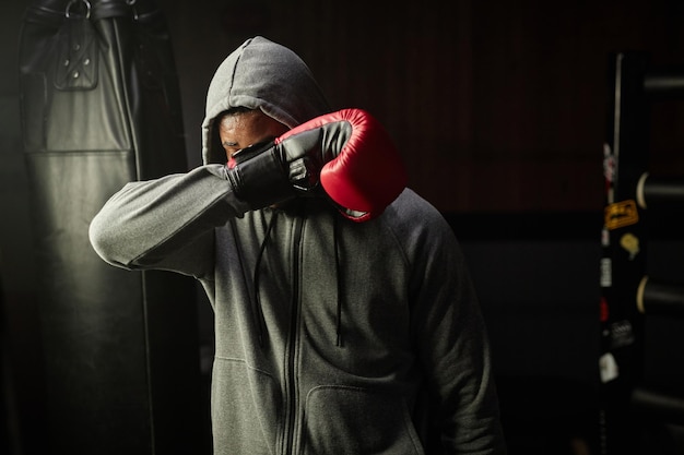 Junger müder afroamerikanischer Boxer im grauen Kapuzenpullover versteckt sein Gesicht hinter einem roten Boxhandschuh, während er ihn im Sportverein vor sich hält