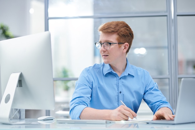 Junger multitasking-büroleiter, der computerbildschirm betrachtet, während er vor laptop sitzt und notizen macht