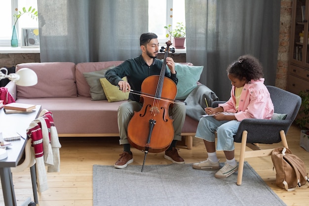 Junger Musiklehrer, der einem biracial Schüler zeigt, wie man Cello spielt, der im Sessel vor ihm sitzt