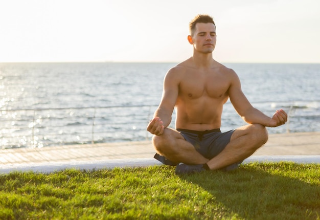 Junger muskulöser Mann mit nacktem Oberkörper meditiert in Lotus-Pose bei Sonnenaufgang am Strand