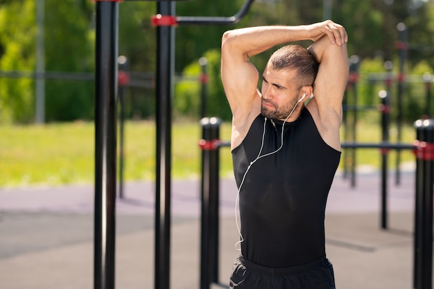 Junger muskulöser Sportler im schwarzen Trainingsanzug, der Übung für Arme beim Stehen auf Sportplatz außerhalb tut