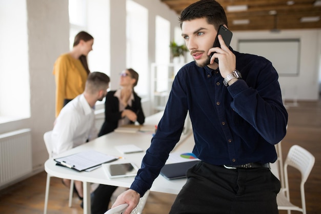 Junger nachdenklicher Geschäftsmann mit Bart im dunklen Hemd, der verträumt zur Seite schaut, während er im Büro mit Kollegen im Hintergrund auf dem Handy spricht