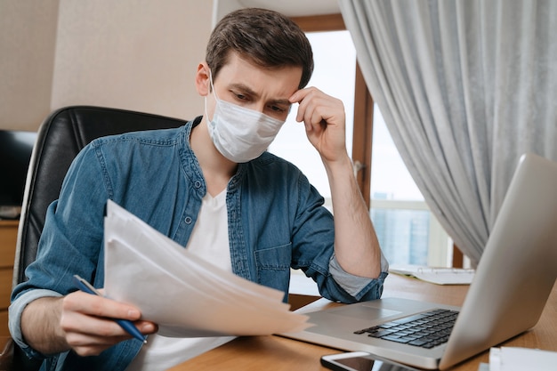 Junger nachdenklicher Mann in schützender Gesichtsmaske, der aufgrund der Coronavirus- oder COVID-19-Quarantäne aus der Ferne studiert und arbeitet