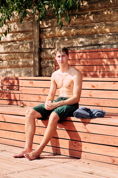 Junger nasser, nackter, männlicher Surfboarder in grünen Shorts, der an einem sonnigen Tag nach hartem Outdoor-Training auf einer Holzbank vor der Kamera sitzt
