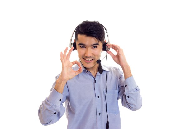 Junger netter Mann mit dunklen Haaren im blauen Hemd mit schwarzen Kopfhörern, die im Studio okay zeigen
