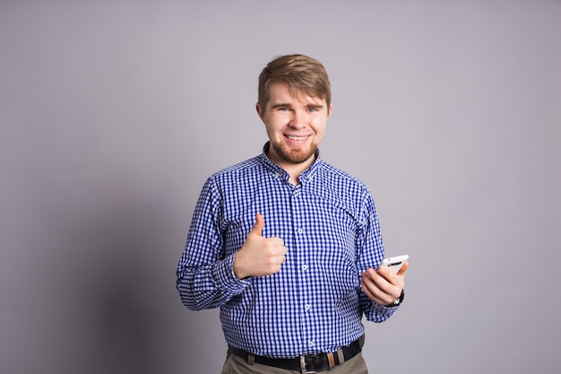 Junger optimistischer Mann mit Handy auf grauer Wand zeigt Daumen hoch mit positiven Emotionen von