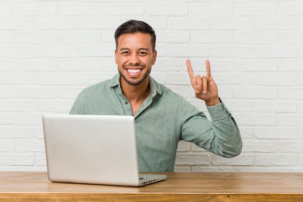 Junger philippinischer Mann, der mit seinem Laptop arbeitet, der eine Hörnergeste als ein Revolutionskonzept zeigt.