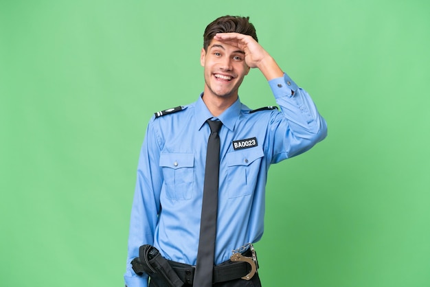 Junger Polizist über isoliertem Hintergrund, der mit der Hand weit wegschaut, um etwas zu sehen