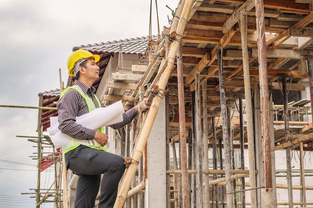 Junger professioneller Ingenieurarbeiter im Schutzhelm und im Blaupausenpapier zur Handarbeit auf der Leiter auf der Baustelle des Hauses