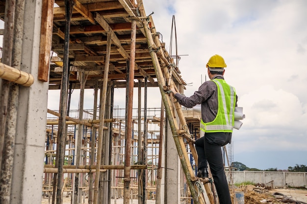 Junger professioneller Ingenieurarbeiter im Schutzhelm und im Blaupausenpapier zur Handarbeit auf der Leiter auf der Baustelle des Hauses