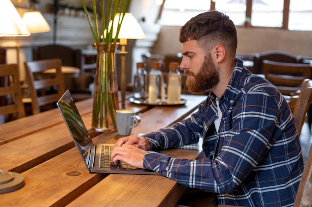 Junger Profi, der auf seinem Laptop mit einer Tasse Kaffee auf dem Tisch im Café oder im Home Office im Internet surft und vom Café-Konzept aus arbeitet