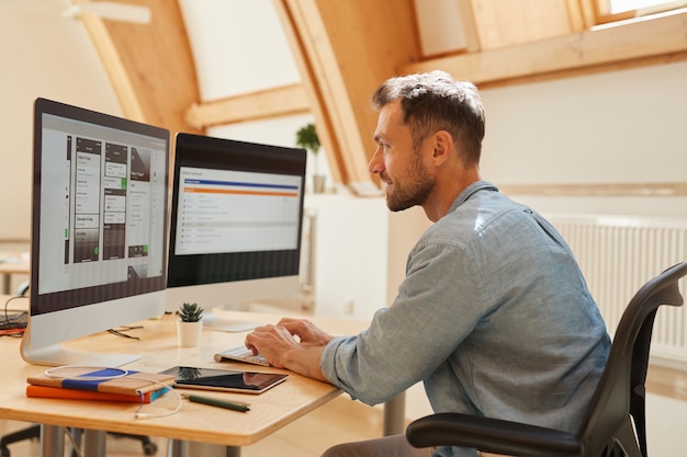 Junger Programmierer, der am Tisch sitzt, der auf Computertastatur tippt und Monitor des Computers betrachtet, den er im Büro arbeitet