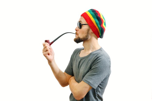 Junger Rasta-Mann in Jamaika-Hut-Sonnenbrille und auf weißem Hintergrund mit Rauchrohr in der Hand