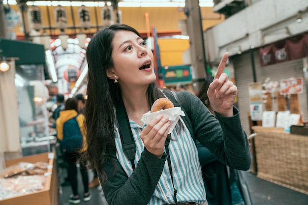 junger reisender, der japanische lokale lebensmittel in osaka probiert. dame tourist zeigt auf den verkäufer und hält einen niedlichen kleinen donut. süßes Straßenessen in Japan.