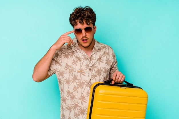 Junger reisender Mann, der einen gelben Koffer auf blauem Hintergrund hält, der eine Enttäuschungsgeste mit Zeigefinger zeigt.