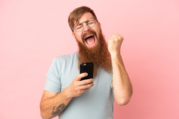 Junger rötlich-kaukasischer Mann isoliert auf rosa Hintergrund mit Telefon in Siegesposition