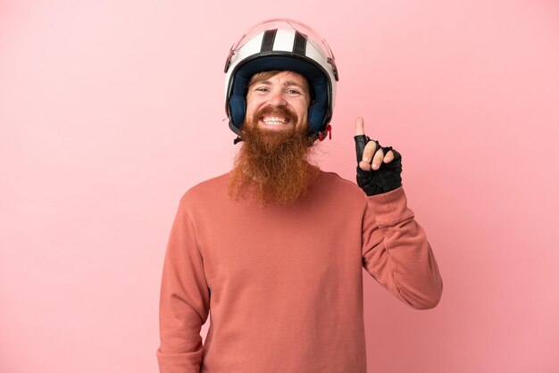 Junger rötlicher kaukasischer Mann mit einem Motorradhelm, der auf rosafarbenem Hintergrund isoliert ist und auf eine großartige Idee zeigt