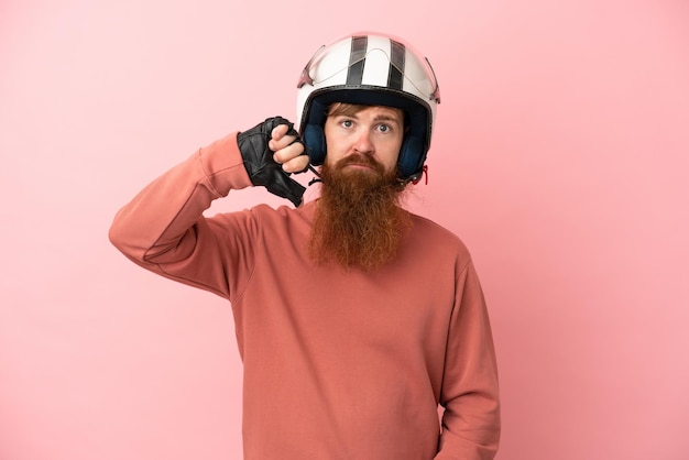 Junger rötlicher kaukasischer Mann mit einem Motorradhelm isoliert auf rosa Hintergrund, der den Daumen nach unten mit negativem Ausdruck zeigt