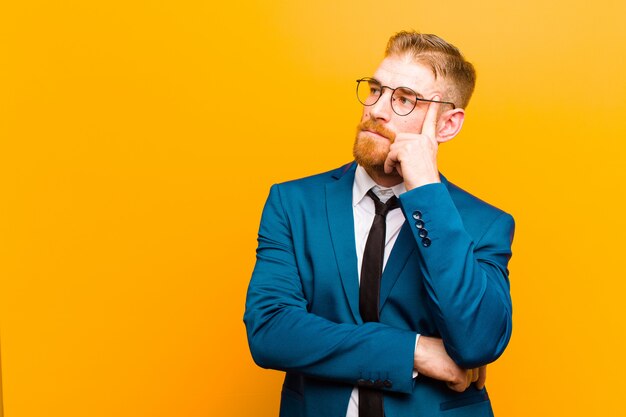 Junger roter Hauptgeschäftsmann mit einem starken Blick, wundernd mit einem zweifelhaften Ausdruck und schauen oben und zur orange Seitenwand