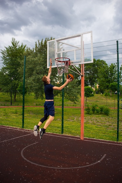 Junger rothaariger Kerl in einem dunkelblauen T-Shirt wirft einen Ball in Bewegung in einen Basketballkorb vor blauem Himmel im Freien