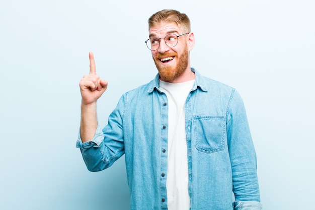 Junger rothaariger Mann, der sich wie ein glückliches und aufgeregtes Genie fühlt, nachdem er eine Idee verwirklicht hat, fröhlich Finger hebend, eureka! über weicher blauer Wand