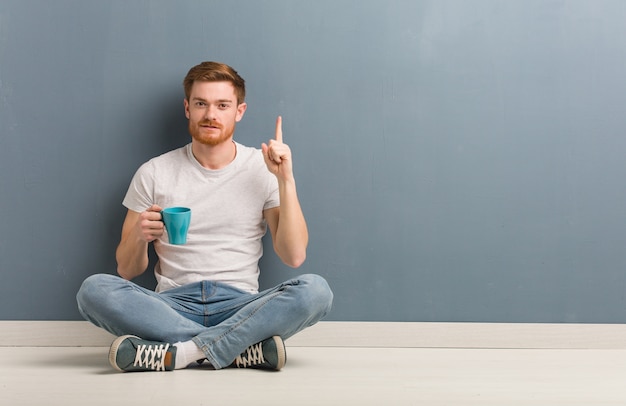 Junger rothaariger Studentenmann, der auf dem Boden sitzt und Nummer eins zeigt. Er hält eine Kaffeetasse in der Hand.