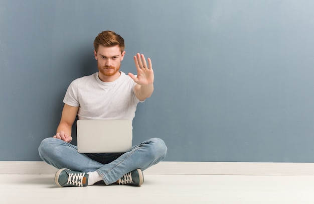 Junger Rothaarigestudentenmann, der auf dem Boden einsetzt Hand in Front sitzt. Er hält einen Laptop in der Hand.