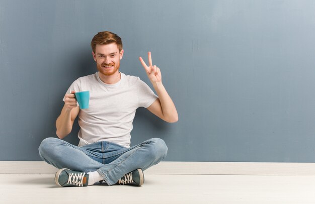 Junger Rothaarigestudentenmann, der auf dem Bodenspaß sitzt und glücklich ist, eine Geste des Sieges tuend. Er hält eine Kaffeetasse in der Hand.