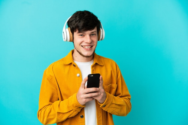 Junger russischer Mann isoliert auf blauem Hintergrund, der Musik mit einem Handy hört und nach vorne schaut