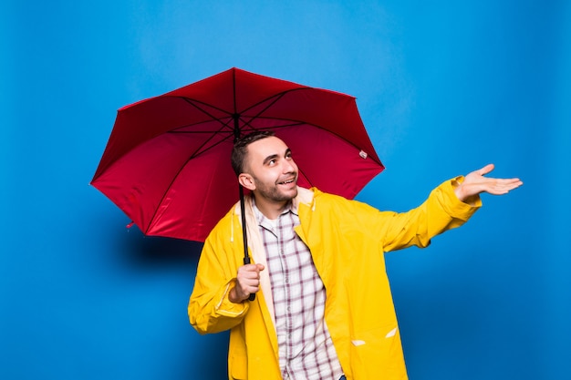 Junger schöner bärtiger Mann im gelben Regenmantel mit rotem Regenschirm lokalisiert über blauem Hintergrund