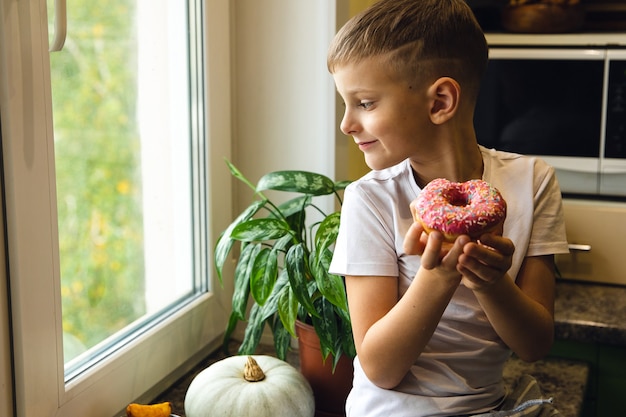 Junger schöner glücklicher und aufgeregter Kinderjunge, der Donut hält und in das Fenster schaut