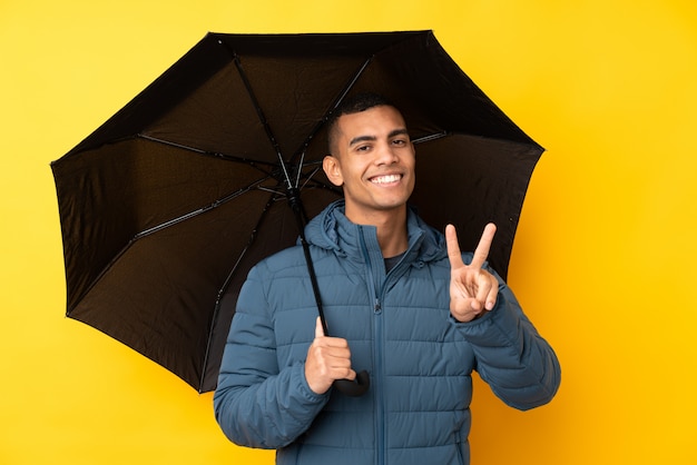 Junger schöner Mann, der einen Regenschirm über der isolierten gelben Wand hält, die lächelt und Siegeszeichen zeigt