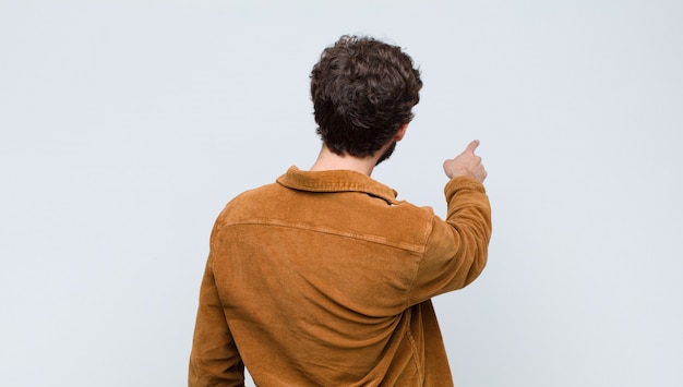 Foto junger schöner mann, der steht und auf objekt auf kopienraum zeigt, rückansicht gegen flache wand