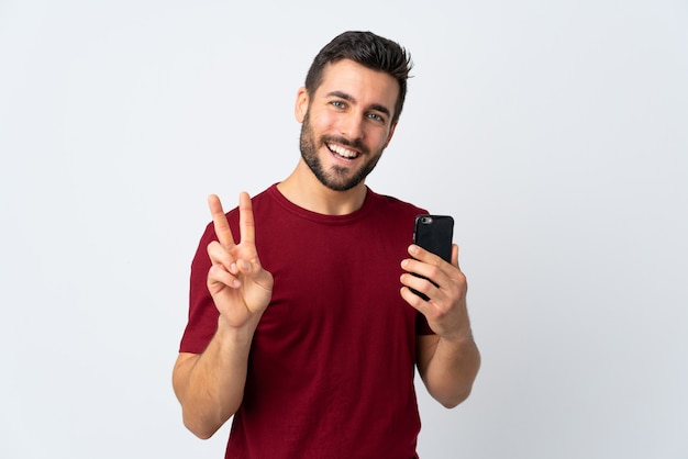 Junger schöner Mann mit Bart unter Verwendung des Mobiltelefons lokalisiert auf der weißen Wand, die lächelt und Siegeszeichen zeigt