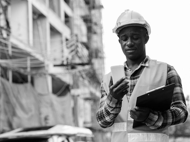 Junger Schwarzafrikanerbauarbeiter, der Zwischenablage hält, während Handy auf der Baustelle verwendet