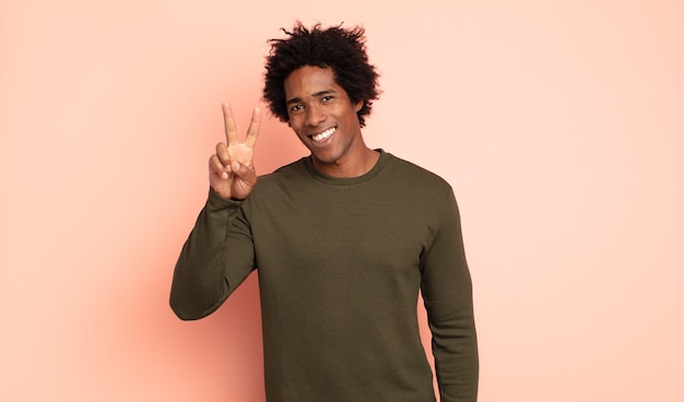 Junger schwarzer Afro-Mann, der lächelt und glücklich, sorglos und positiv schaut, Sieg oder Frieden mit einer Hand gestikulierend