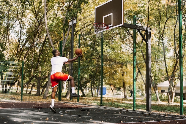 Junger schwarzer lächelnder Mann, der bei Sonnenaufgang Basketball auf dem Platz spielt, Morgensport. Outdoor-Porträt.