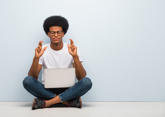 Junger schwarzer Mann, der auf dem Boden mit den Fingern einer Laptopüberfahrt für das Haben des Glücks sitzt