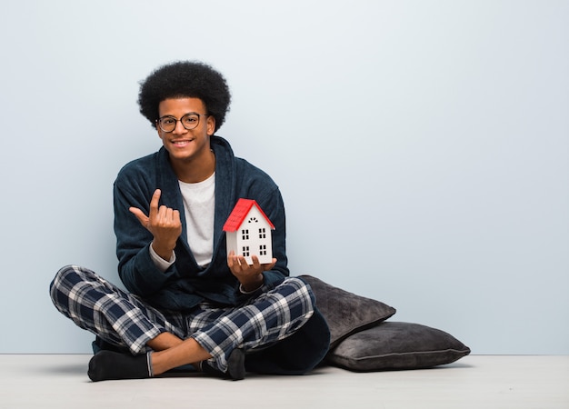 Junger schwarzer Mann, der ein Hausmodell sitzt auf dem Boden einlädt zu kommen hält