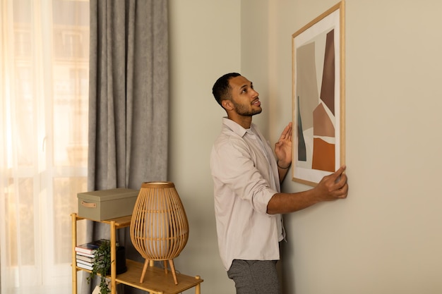 Junger schwarzer Mann, der einen Bilderrahmen aufhängt, der ein Gemälde an der Wand aufhängt. Innen- und Inneneinrichtung