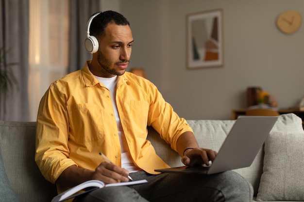 Junger schwarzer Mann mit Kopfhörern, der eine Online-Geschäftskonferenz oder ein Webinar auf dem Laptop hat und sich zu Hause Notizen macht